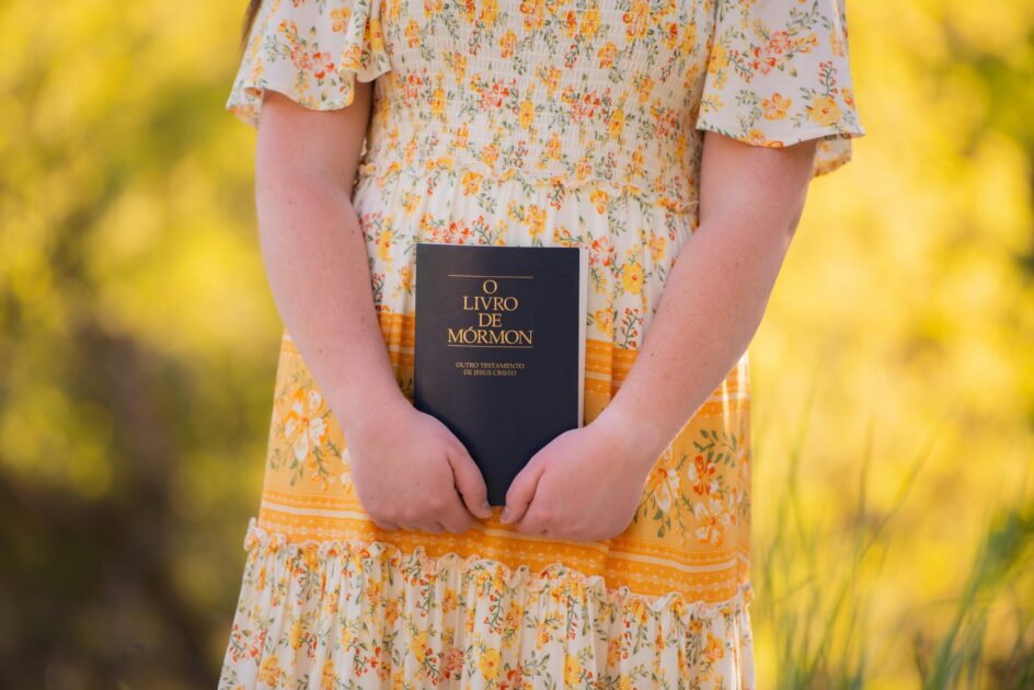A child holds a Book of Mormon in a floral dress, set against a bright outdoor background.