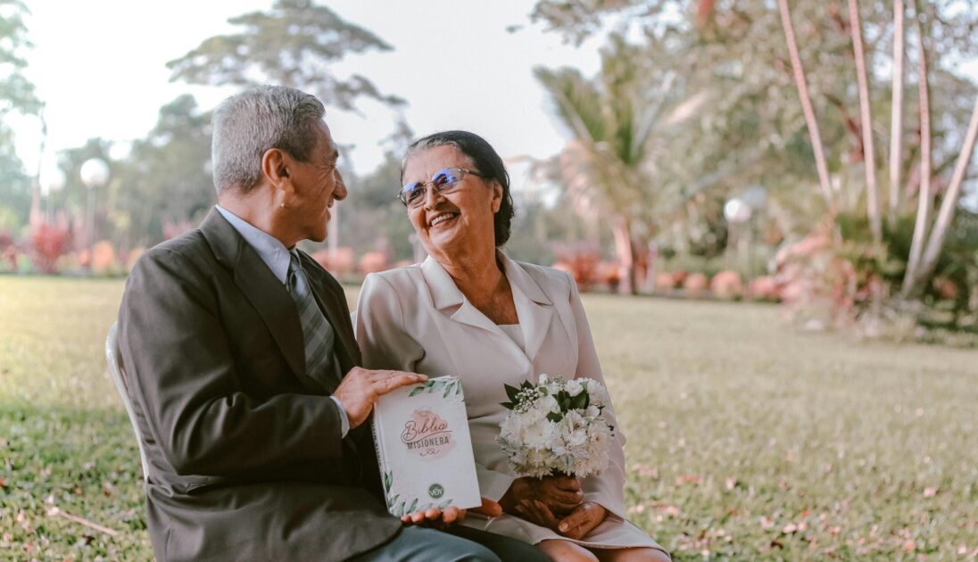 Smiling elderly couple holding flowers, sharing a moment in a sunny park.