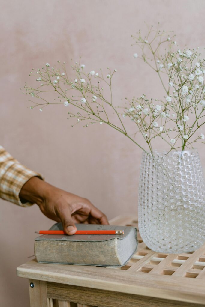 A hand reaches for a vintage book beside a vase of white flowers on a wooden table.