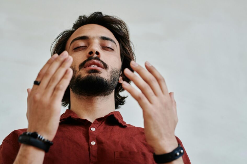 A man with a beard wearing a red shirt meditates with eyes closed in a serene atmosphere.