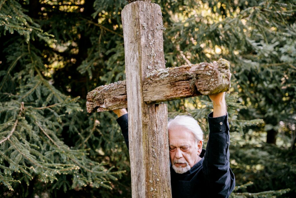 An emotional elderly man embraces a rustic wooden cross in a serene forest setting.